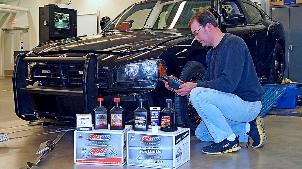 Technician holds an AMSOIL bottle beside a black Dodge Charger police car on a dyno, with AMSOIL products and cases on the floor