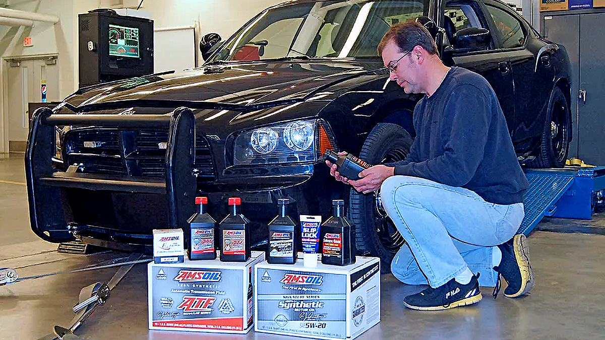 Technician holds an AMSOIL bottle beside a black Dodge Charger police car on a dyno, with AMSOIL products and cases on the floor