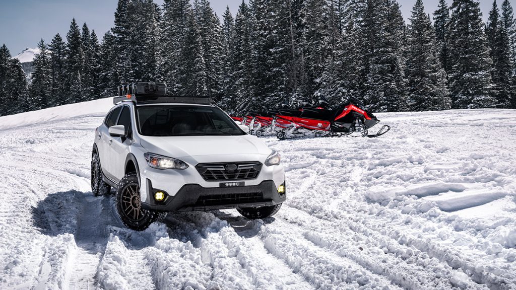 A Suburu Crosstrek is parked in snow with snowmobiles in the background.