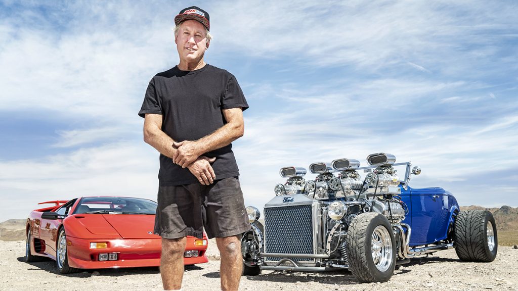 Gordon Tronson stands between a red supercar and a supercharged hot rod he built